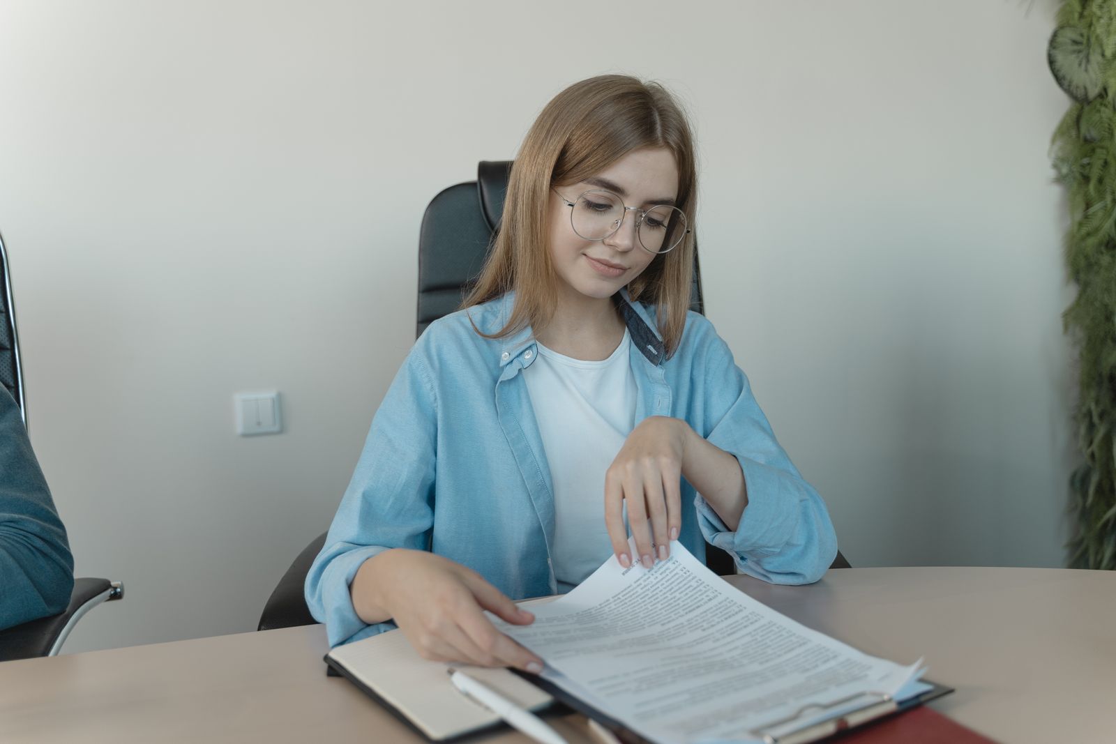 Uma mulher de óculos folheia documentos impressos em uma prancheta, sentada à mesa.
