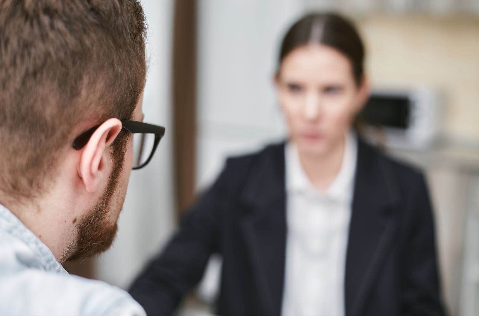Um homem de barba e óculos, visto de costas, de frente para uma mulher de blazer desfocada.