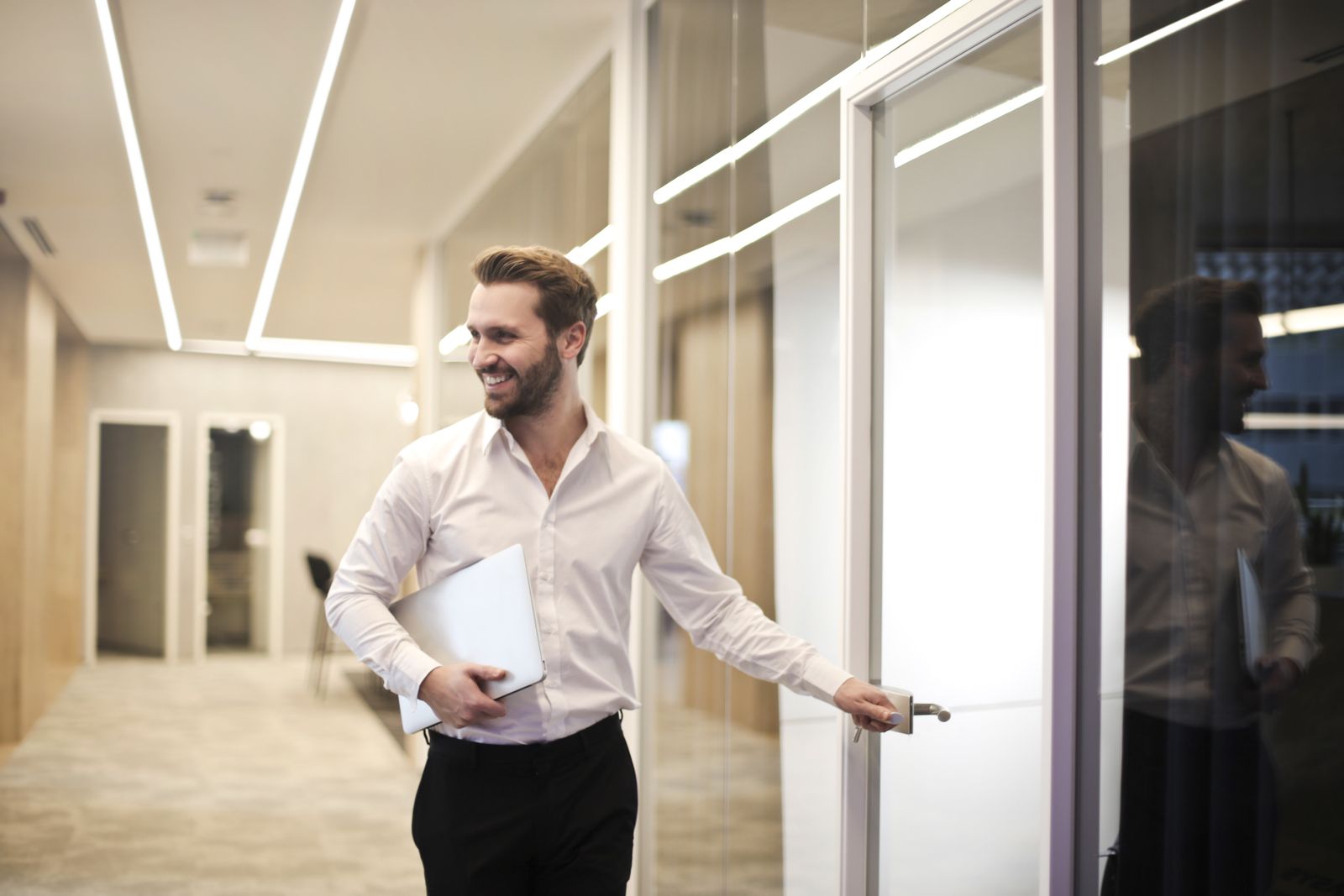 Um homem sorridente carregando um notebook abre uma porta de vidro em um corredor de escritório.